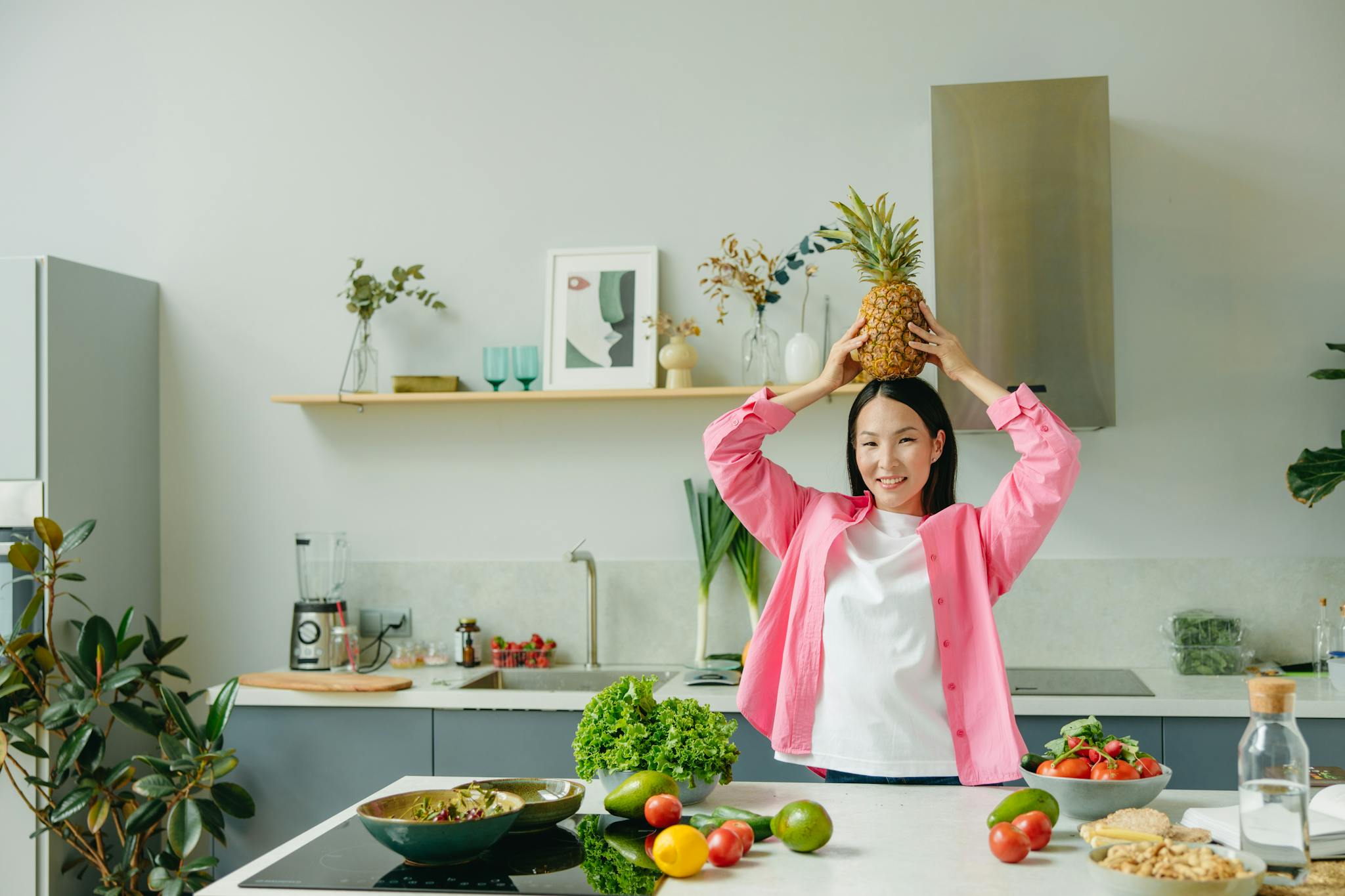 Asian woman in pink overshirt holding a pineapple in a modern kitchen with fresh produce and cheerful ambiance.