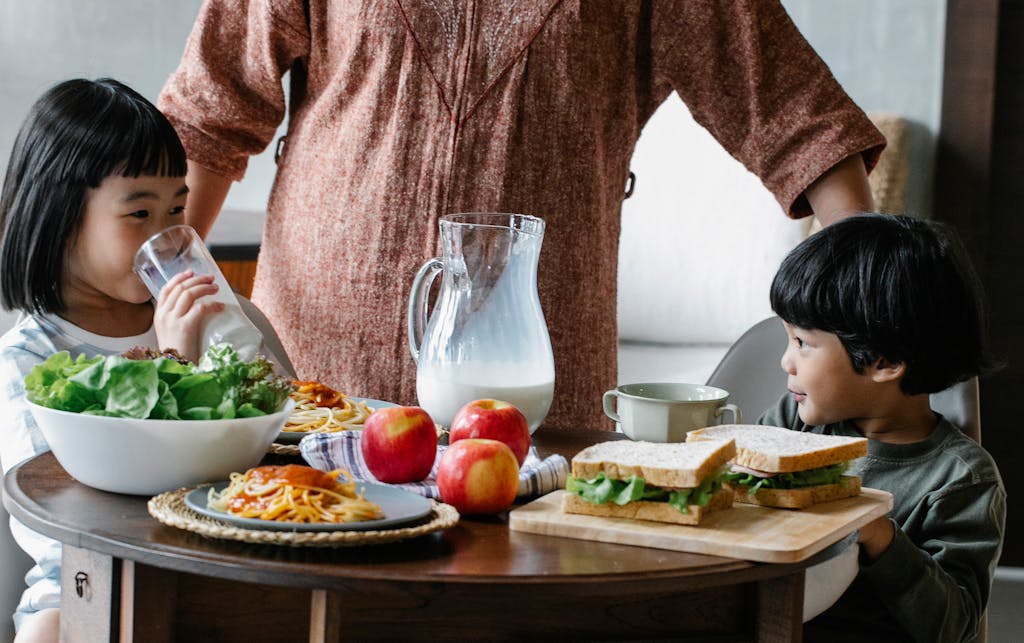 Unrecognizable female in casual clothes standing at served table with sandwiches and milk near Asian boy and girl during breakfast in kitchen